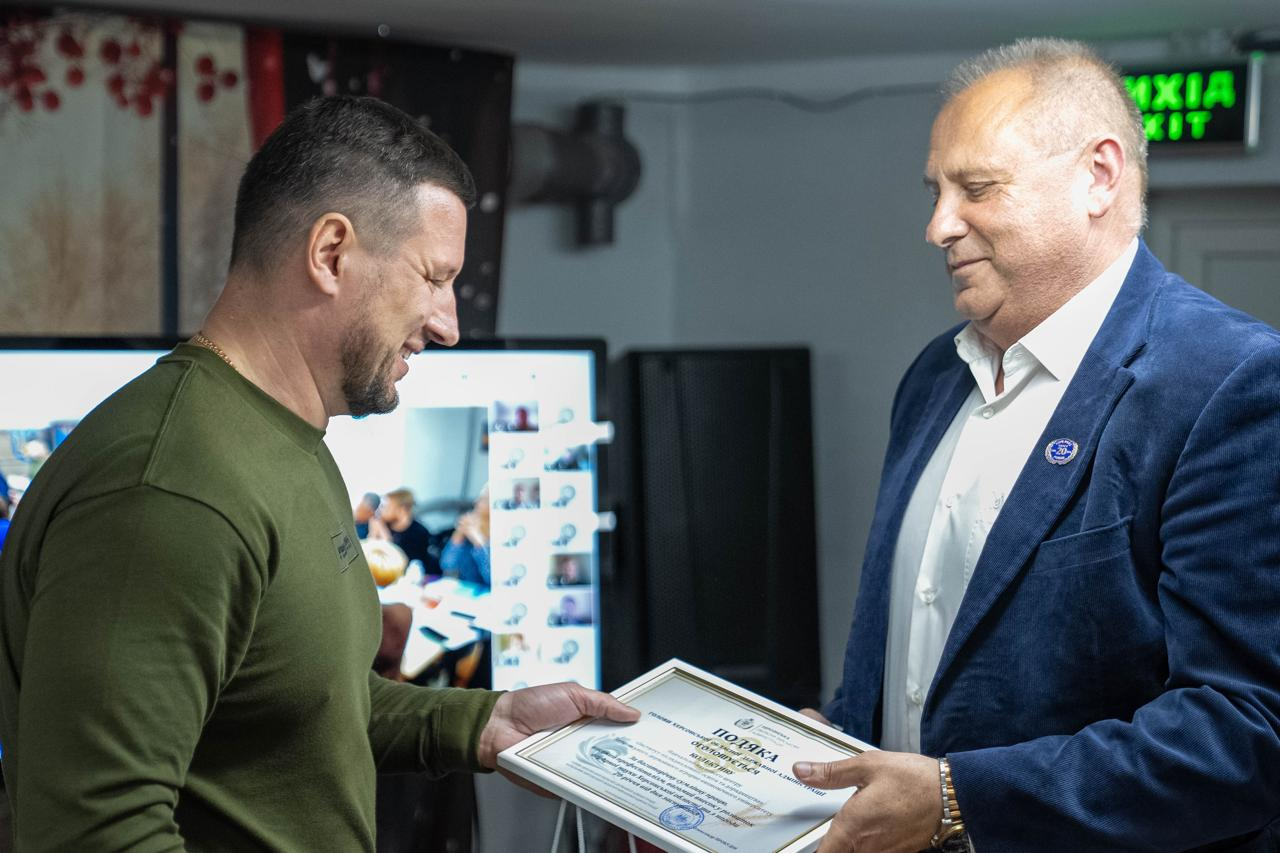 Урочисте вручення відзнаки, де чоловік у військовому одязі хакі передає офіційну грамоту-подяку в рамці чоловікові у синьому піджаку та білій сорочці.