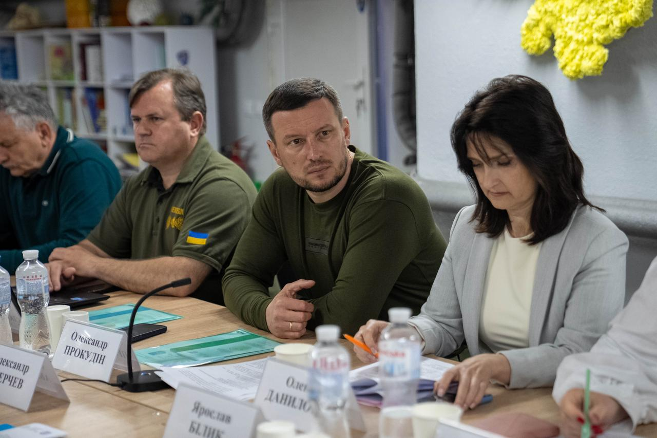 Нарада в робочому кабінеті, де учасники сидять за столом із документами та табличками. У центрі уваги — чоловік у військовому одязі хакі, який уважно слухає колег, а поруч із ним сидить жінка у світлому жакеті.