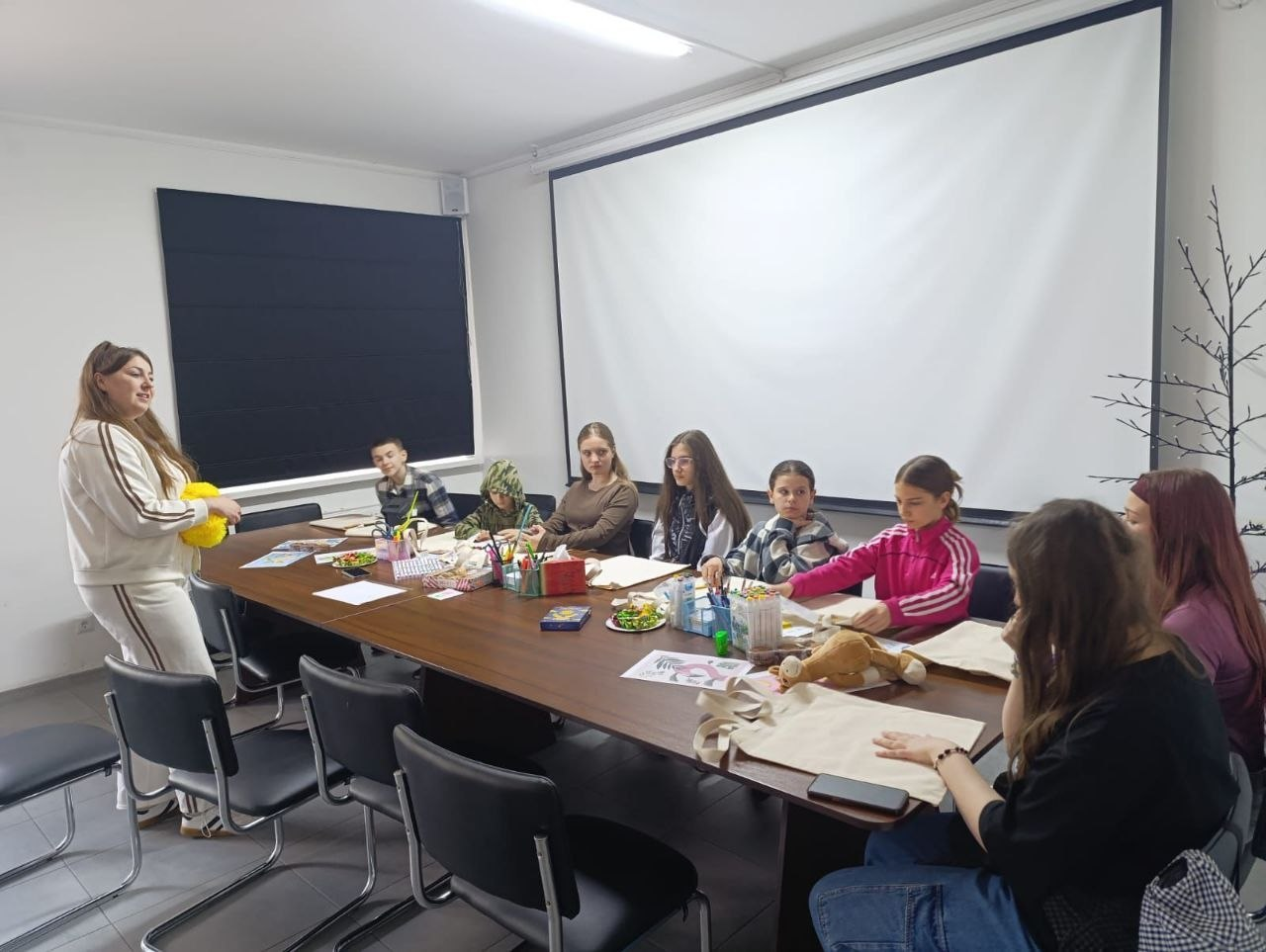 Творче заняття у навчальній аудиторії. Ведуча у світлому костюмі спілкується з групою дітей та молоді, які сидять навколо великого столу з екосумками та канцелярським приладдям, а позаду них розміщено великий білий екран.