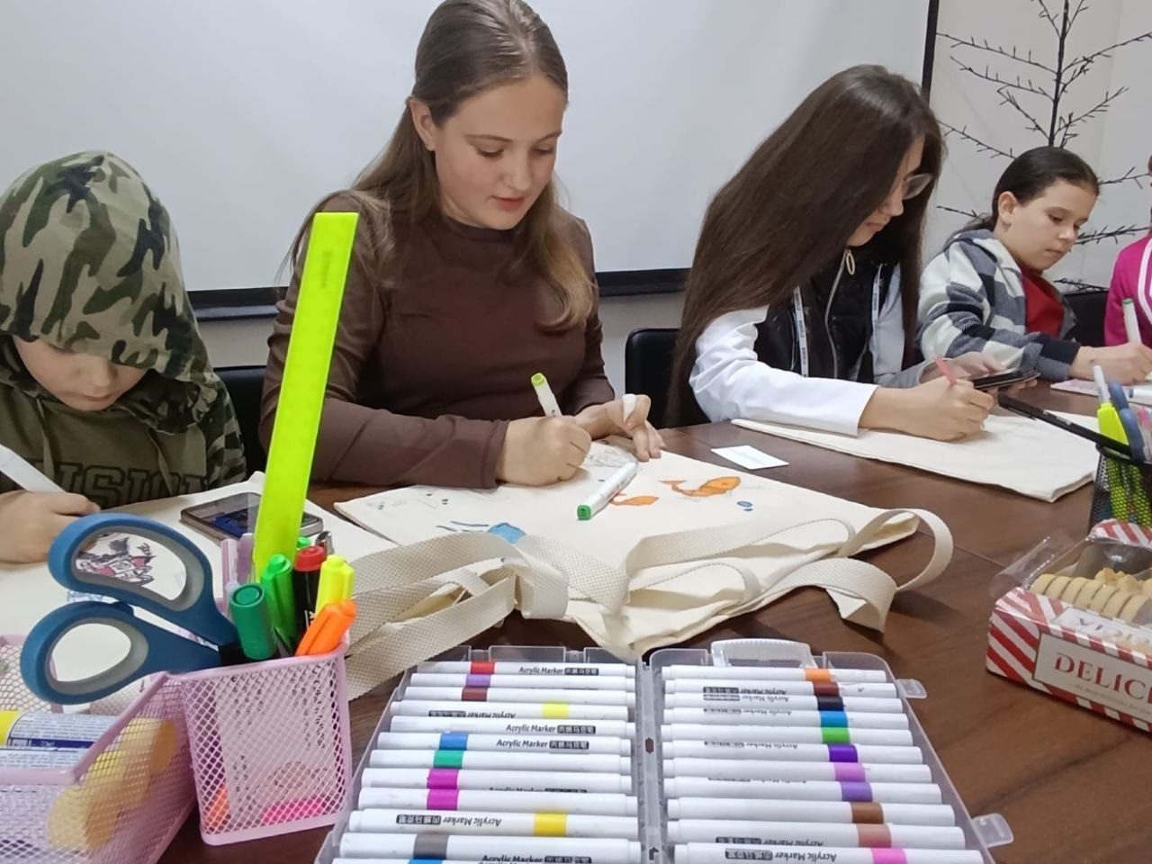 Творче заняття, на якому діти та молодь сидять за столом і розмальовують маркерами світлі екосумки. На передньому плані зблизька видно великий набір різнокольорових акрилових маркерів та підставку з канцелярією.