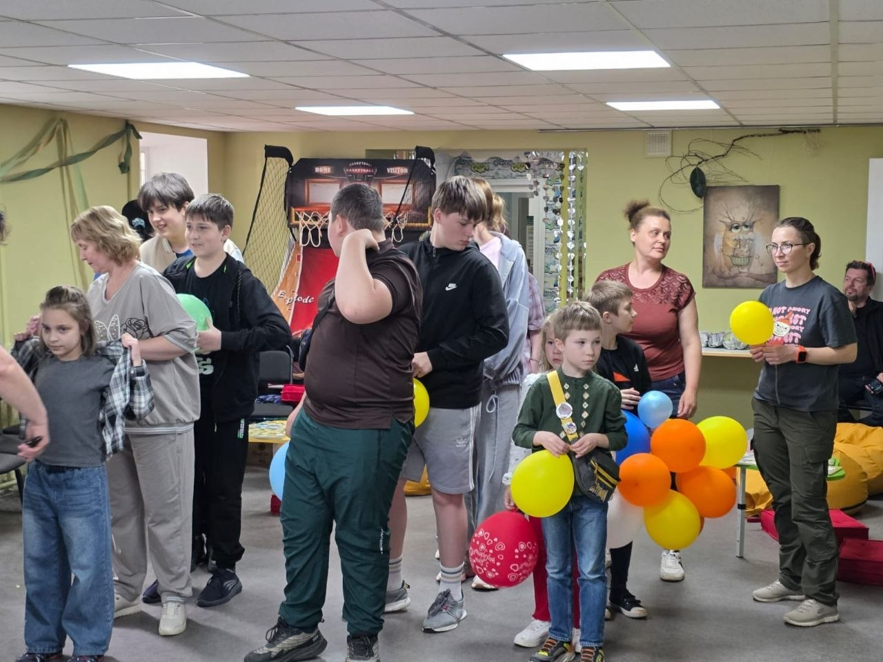 Група дітей та дорослих під час розважального заходу чи гри у приміщенні. Учасники стоять у залі, дехто тримає в руках різнокольорові повітряні кульки, а на задньому плані видно дитяче баскетбольне кільце та декор на стінах.
