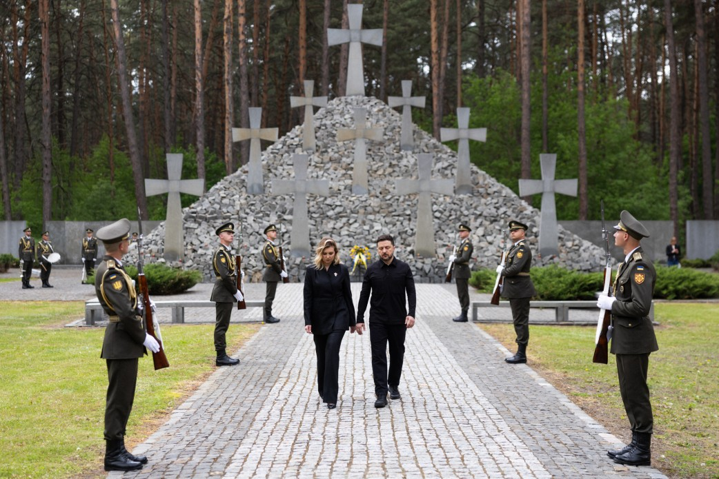 Чоловік та жінка у чорному вбранні йдуть брукованою доріжкою від меморіалу з великими кам'яними хрестами на тлі хвойного лісу. Вздовж доріжки вишикувалася почесна варта військовослужбовців у парадній формі.