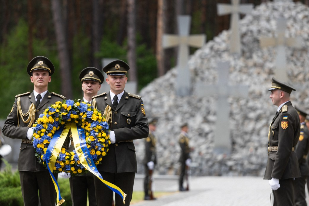 Військовослужбовці у парадній формі несуть великий синьо-жовтий вінок під час офіційної церемонії вшанування пам'яті. На розмитому задньому плані видно кам'яний меморіал із хрестами.