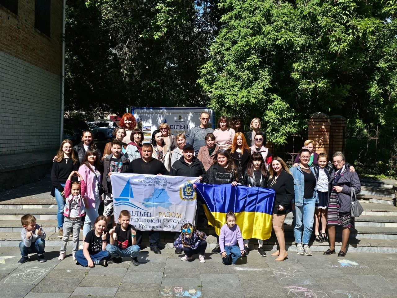 Велика група дітей та дорослих позує для спільного фото просто неба на тлі цегляної будівлі та зелених дерев. Учасники тримають великий синьо-жовтий прапор України та білий банер із патріотичними написами й малюнком корабля.
