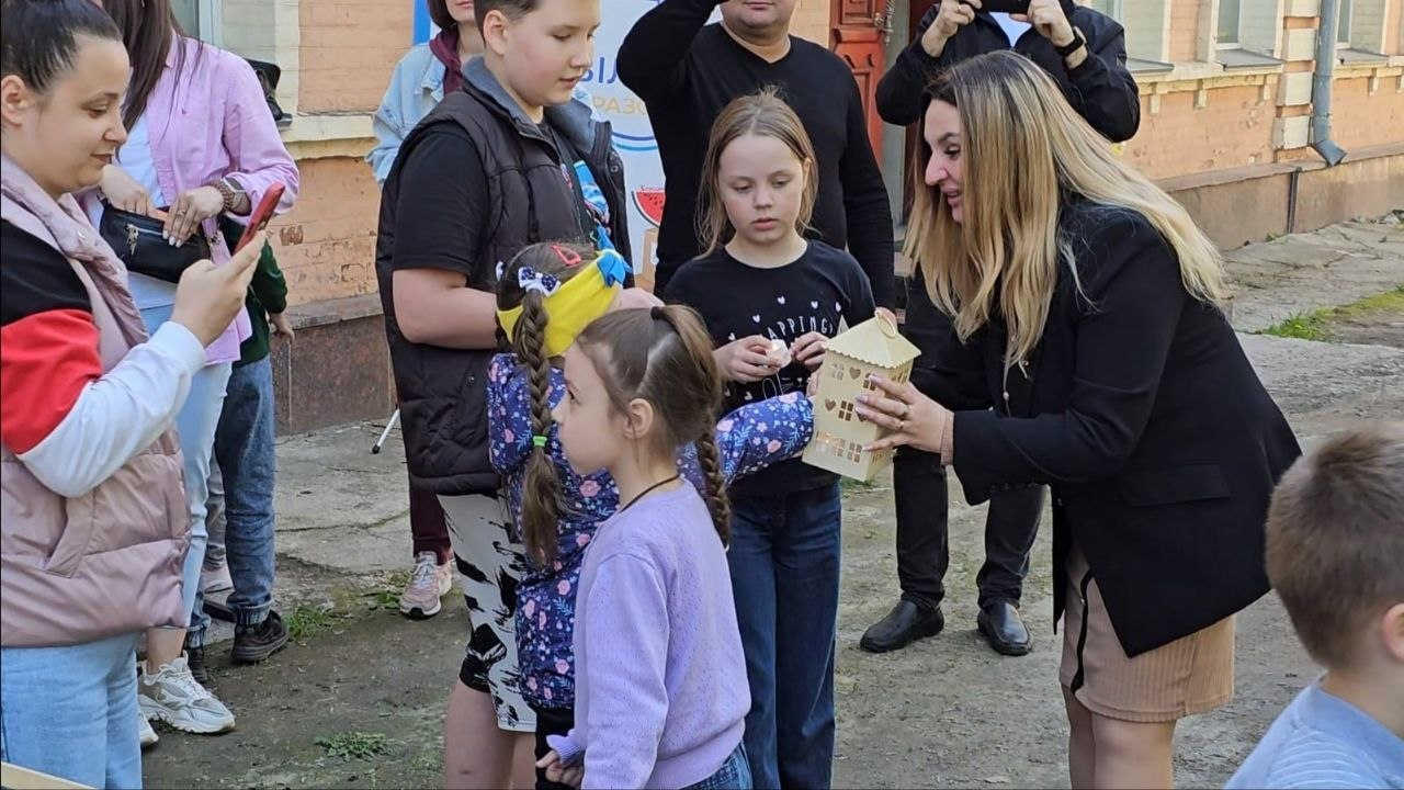 Група дітей та дорослих на подвір'ї просто неба під час заходу. Жінка у чорному жакеті передає дівчинці декоративний картонний будиночок, а інші присутні поруч спостерігають за процесом та знімають подію на телефони.