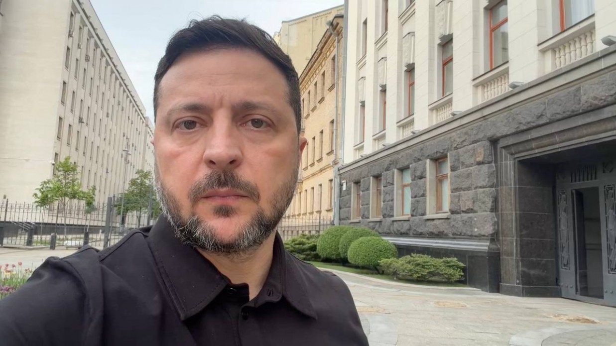 Чоловік у чорній сорочці робить селфі-знімок просто неба на тлі великої урядової будівлі з сірим кам'яним цоколем та декоративними кущами.