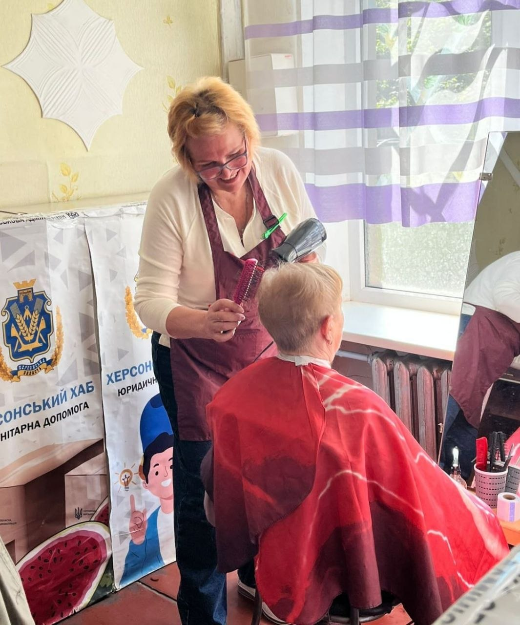 Жінка-перукар у фартуху робить укладку волосся іншій жінці за допомогою фену та круглої щітки у приміщенні біля вікна.