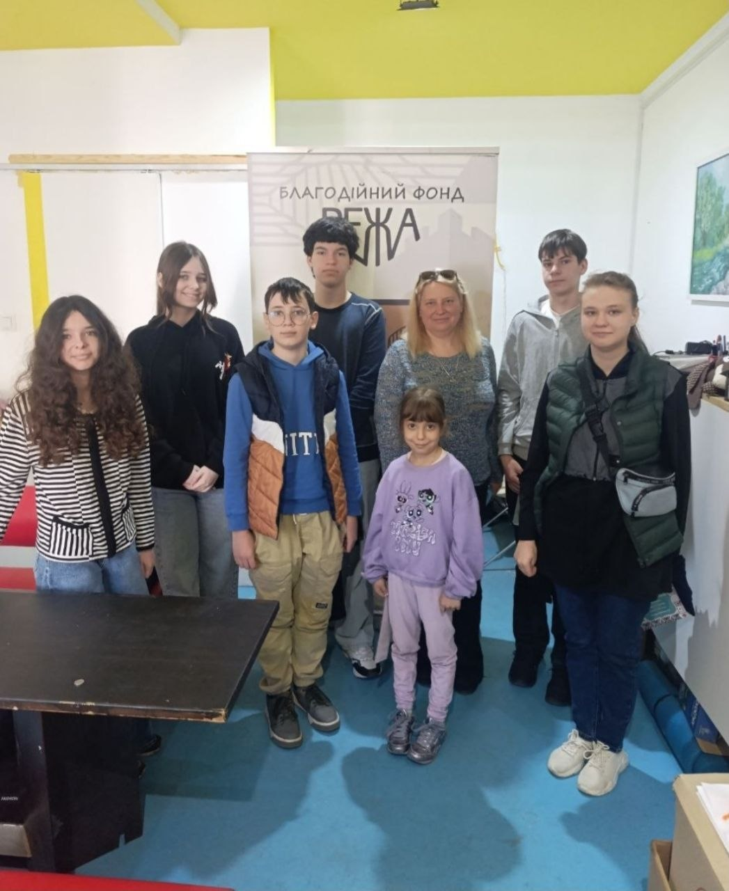 Група з семи дітей різного віку та однієї дорослої жінки стоїть у приміщенні на тлі банера з написом «Благодійний фонд 