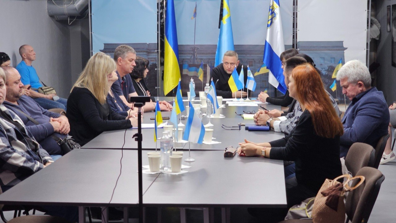 Тематична зустріч представників влади та громадськості за довгим столом у конференц-залі, де учасники сидять один навпроти одного, а на фоні та столі встановлено державні прапори України та прапори Херсона.