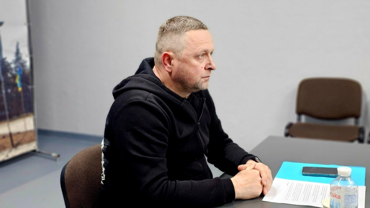 Чоловік у чорному худі сидить за столом у профілі під час офіційної зустрічі, склавши руки перед собою, поруч із документами, телефоном та пляшкою води.