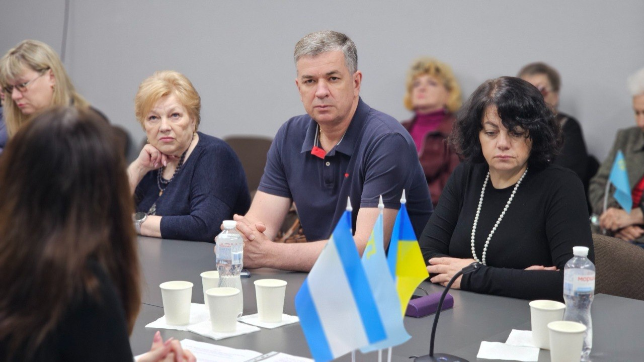 Учасники тематичної зустрічі сидять за столом у конференц-залі, слухаючи спікера, а перед ними на столі встановлено настільні прапорці України та міста Херсона, паперові стаканчики та пляшки з водою.