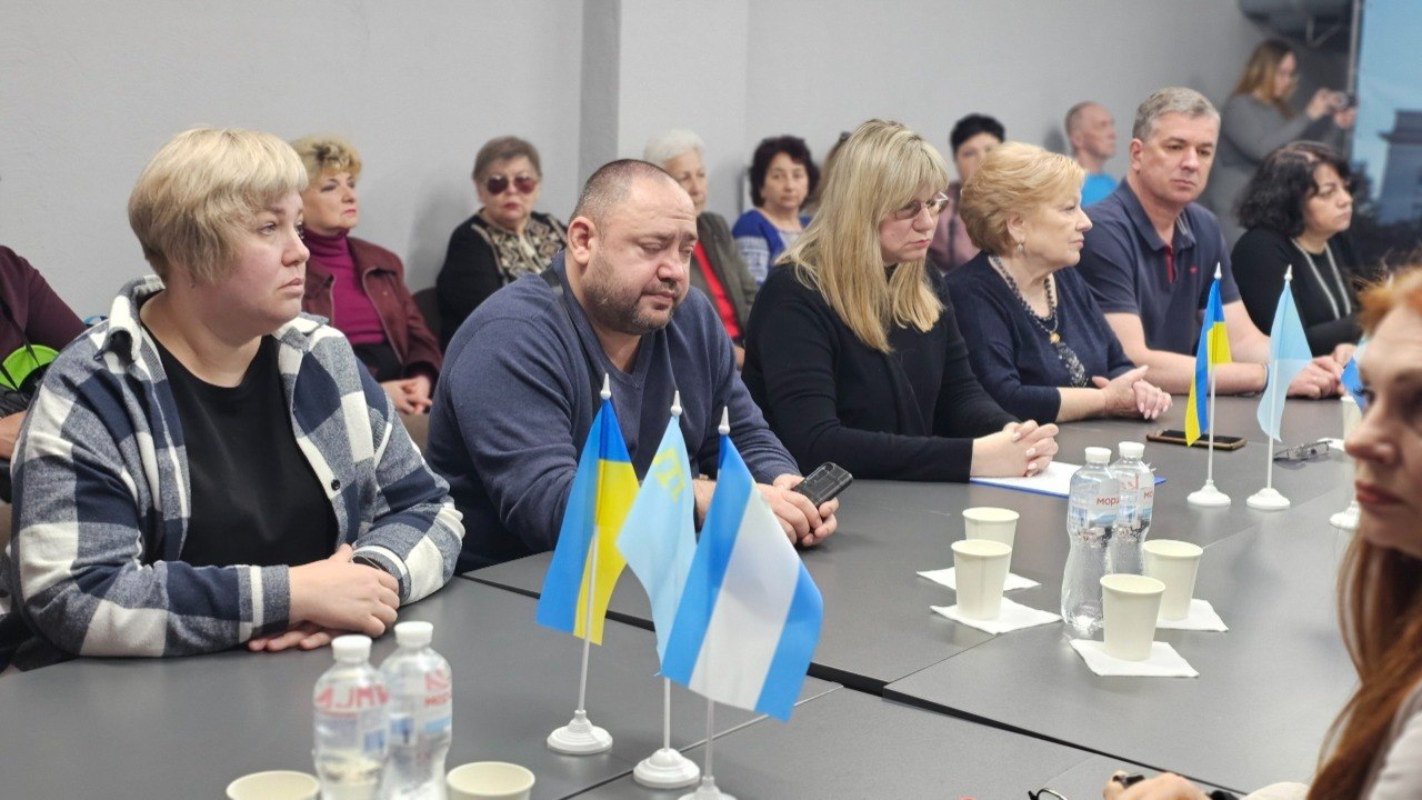 Учасники тематичної зустрічі сидять уздовж довгого столу в конференційній залі, на якому розставлені маленькі прапори України та міста Херсона, пляшки з водою та паперові стаканчики.