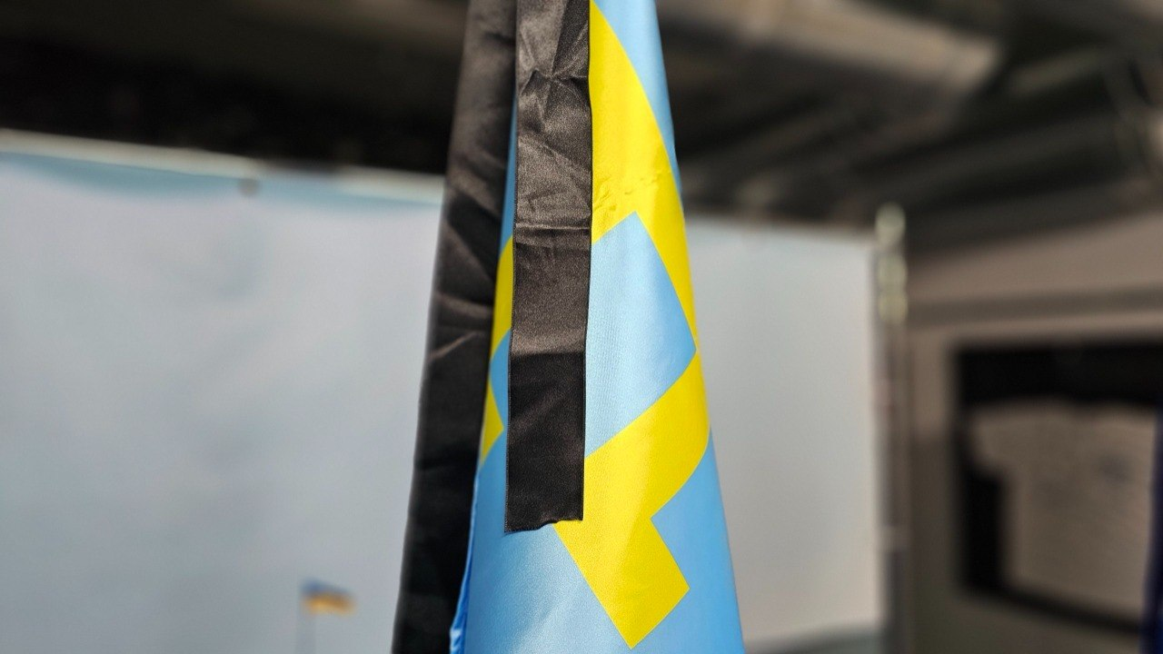 Крупний план прапора міста Херсона із прив'язаною до нього чорною жалобною стрічкою у приміщенні на розмитому тлі банерів.