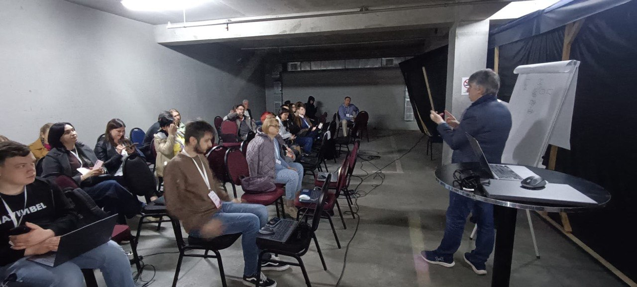 Спікер виступає перед аудиторією під час лекції або тренінгу, що проходить у підземному приміщенні чи укритті з бетонними стінами, використовуючи ноутбук та фліпчарт.