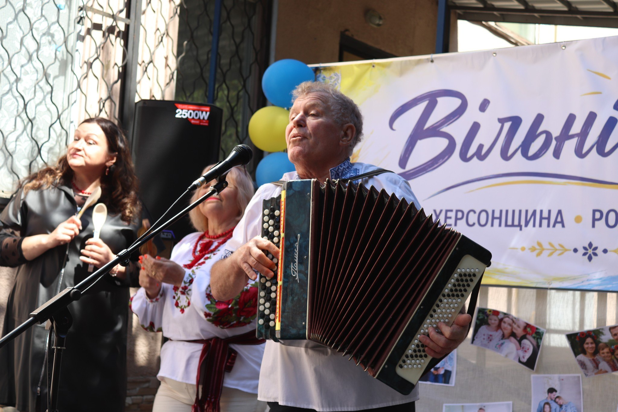 Чоловік у вишиванці грає на баяні та співає у мікрофон на вуличній сцені поруч із двома жінками на тлі святкового банера «Вільні разом».