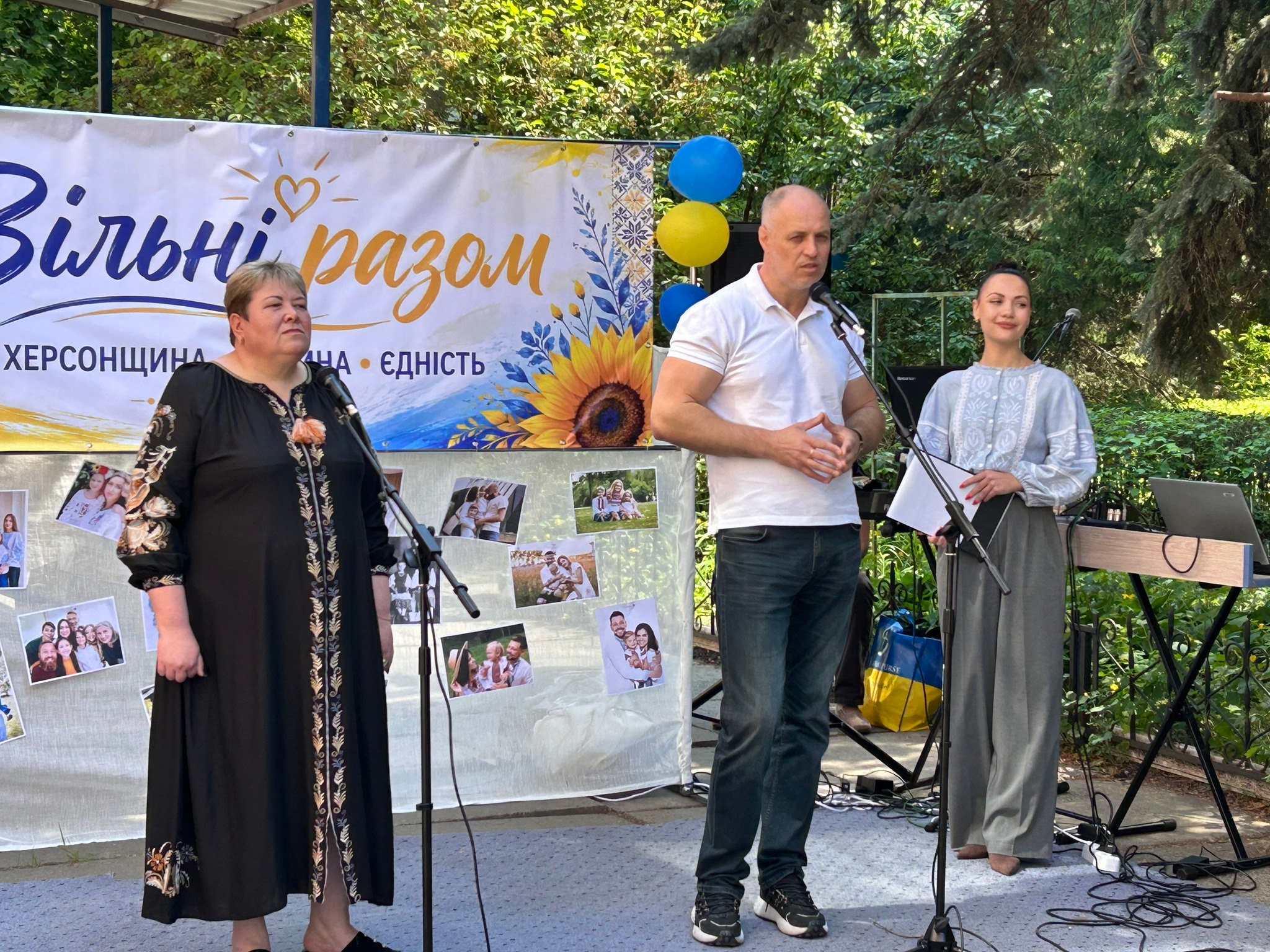 Чоловік та дві жінки виступають із промовами біля мікрофонів на вуличній сцені на тлі банера з написом «Вільні разом. Херсонщина • Родина • Єдність» та фотокартками.