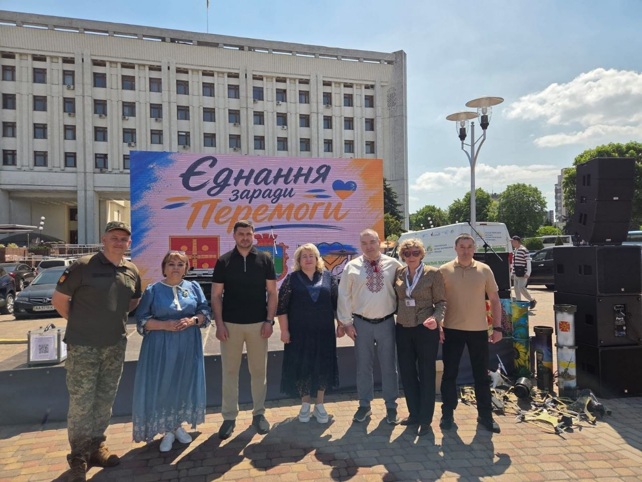Група із семи людей позує на площі перед адміністративною будівлею на тлі банера «Єднання заради Перемоги». Вони одягнені так (зліва направо): чоловік у військовій формі, жінка в блакитній сукні, чоловік у чорному поло, жінка в темній сукні, чоловік у білій вишиванці, жінка в кавовій блузці та чоловік у бежевому поло.