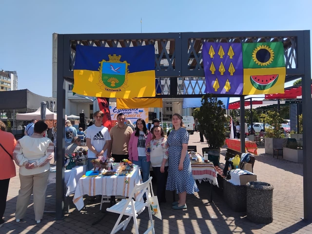 Група людей стоїть на вуличній локації біля виставкового столу під металевим навісом, на якому закріплені прапори з гербами Чаплинки та Долматівської громади.