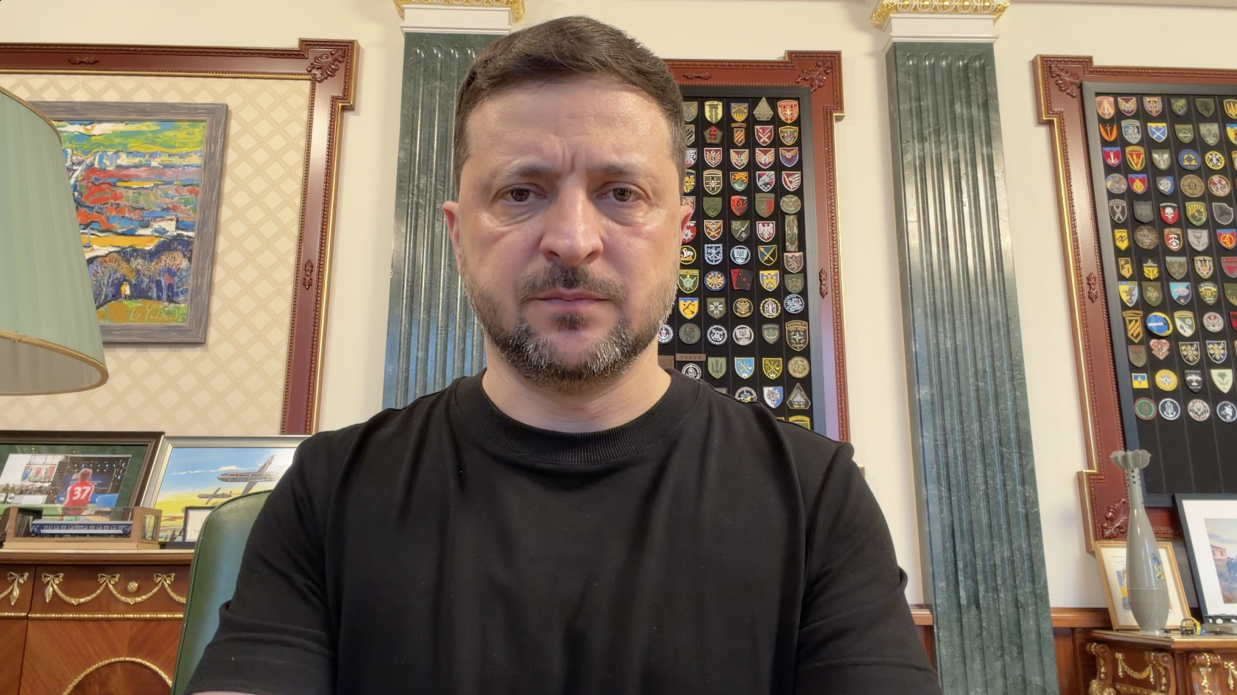 Чоловік у чорній футболці дивиться прямо в кадр, сидячи в робочому кабінеті, де на задньому плані видно стіни з великою колекцією військових шевронів у рамках, картини та елементи класичного інтер'єру.