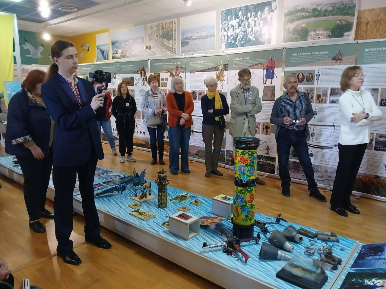 Група людей стоїть у музейній залі біля виставкових стендів та довгого столу, на якому розкладено різноманітні експонати, зокрема військові артефакти й розписаний тубус. Один із присутніх чоловіків тримає камеру та знімає захід.
