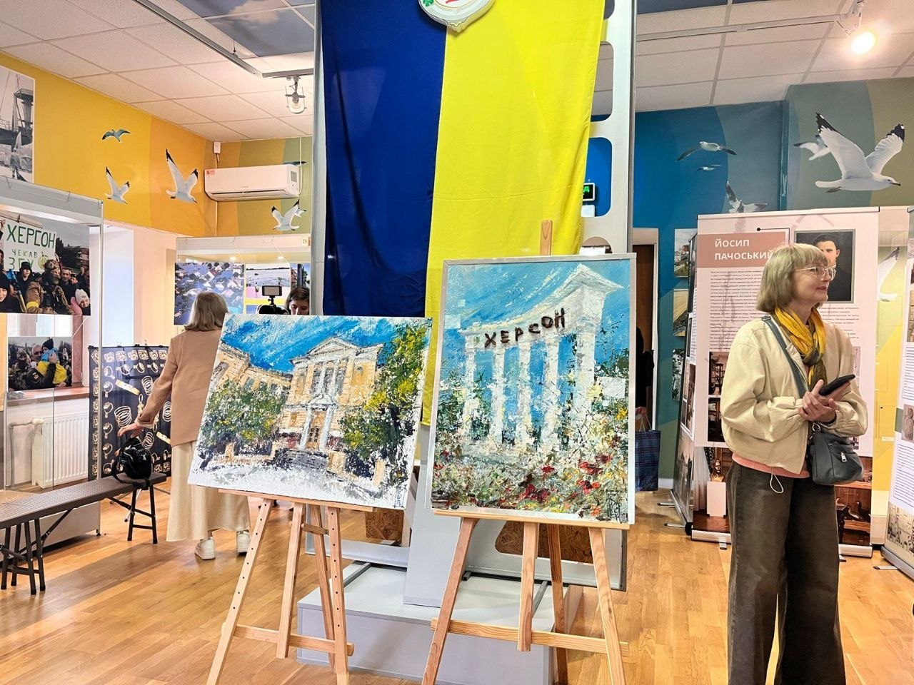 Дві картини на дерев'яних мольбертах, що зображують архітектурні пам'ятки Херсона, стоять на тлі великого синьо-жовтого прапора у виставковій залі музею. На задньому плані та праворуч видно відвідувачів та інформаційні стенди.