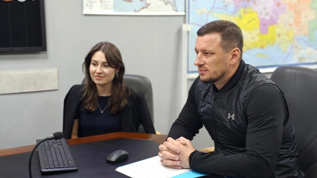 Чоловік та жінка сидять за робочим столом у кабінеті на тлі географічних карт на стіні. Перед чоловіком лежать документи, а на столі ліворуч розташовані клавіатура, комп'ютерна миша та мікрофон.