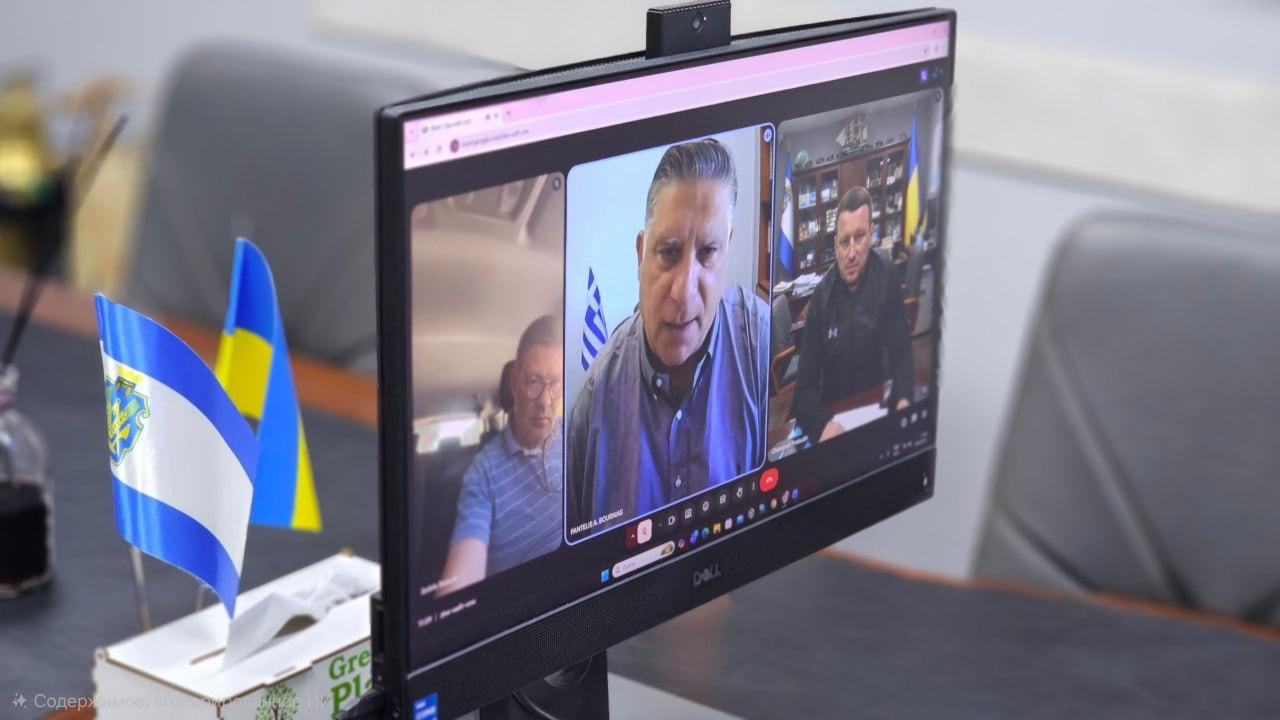 Комп'ютерний монітор відображає онлайн-відеоконференцію за участю трьох осіб, знятий збоку на робочому столі. Ліворуч на столі перед екраном стоять два маленькі настільні прапорці — Державний Прапор України та прапор із гербом міста.
