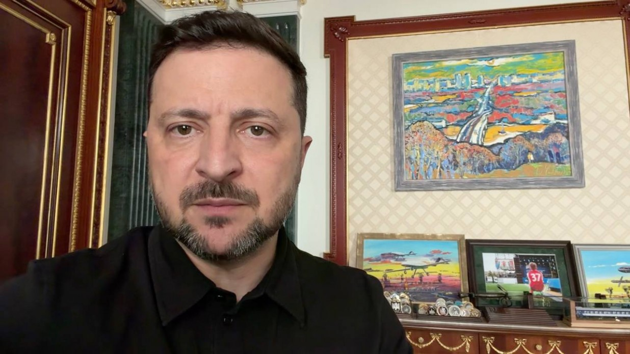 Чоловік у чорній сорочці знятий крупним планом на тлі кабінету з класичним інтер'єром. На стіні позаду нього висить яскрава картина із зображенням пейзажу, а нижче на полиці розставлені інші невеликі картини, фотографії у рамках та сувеніри.