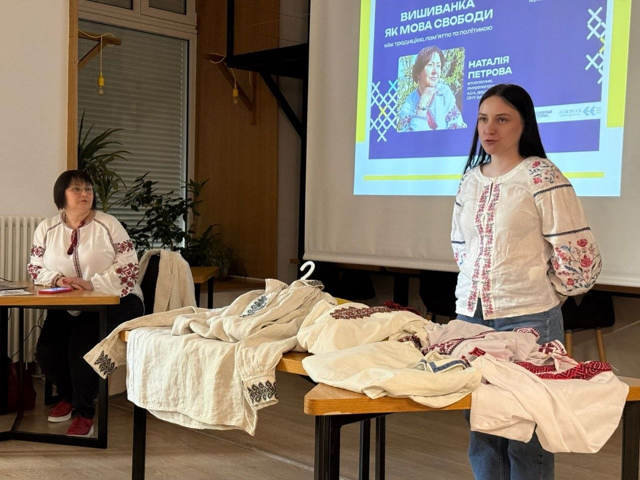 Фото з лекції-дискусії, присвяченої українській вишиванці. Жінка у вишитій сорочці виступає на тлі екрана зі слайдом «Вишиванка як мова свободи», а на столі перед нею розкладено зразки традиційного вишитого одягу.