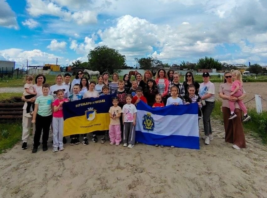 Групове фото дорослих та дітей просто неба на тлі сільського пейзажу та коней. Учасники зустрічі стоять разом і тримають перед собою два великі прапори: ліворуч — синьо-жовтий прапор Верхньорогачицької територіальної громади, праворуч — біло-синій прапор Херсонської області.
