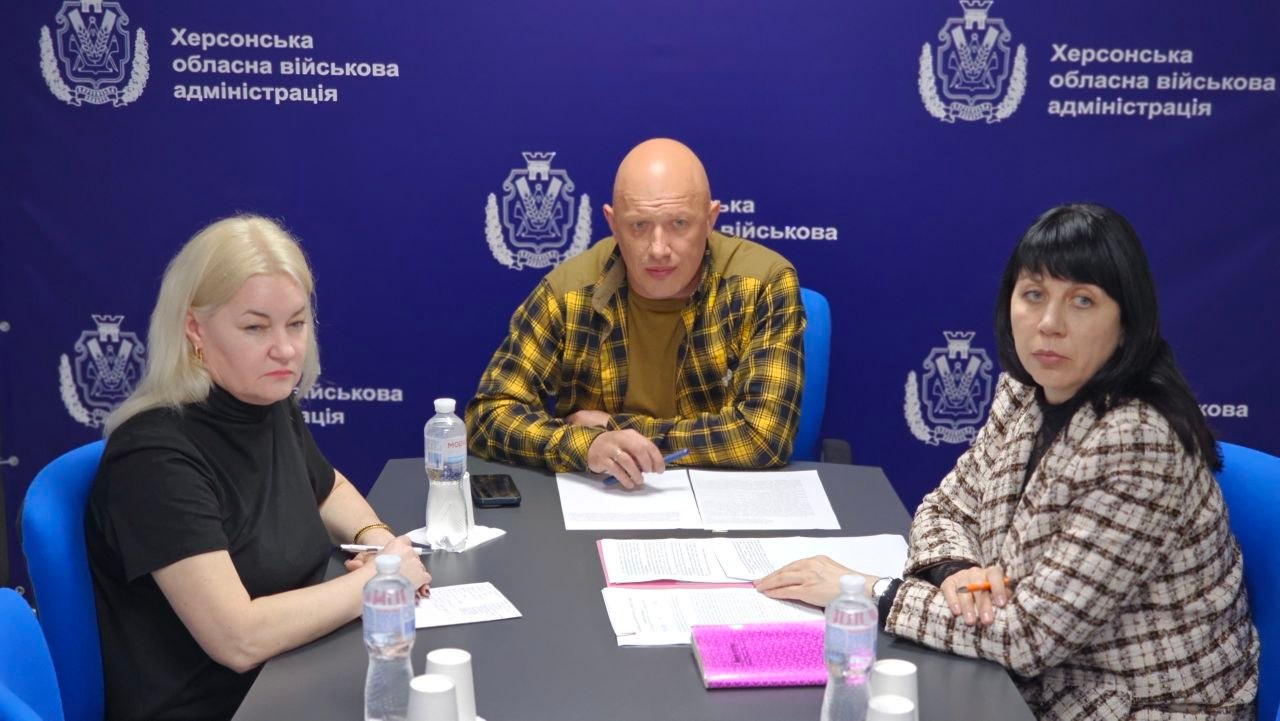 Три особи — дві жінки та чоловік у центрі — сидять за столом під час робочої наради чи офіційного виступу. Перед ними розкладені документи, канцелярське приладдя та пляшки з водою, а на синьому тлі позаду розміщено офіційні герби та написи установи.