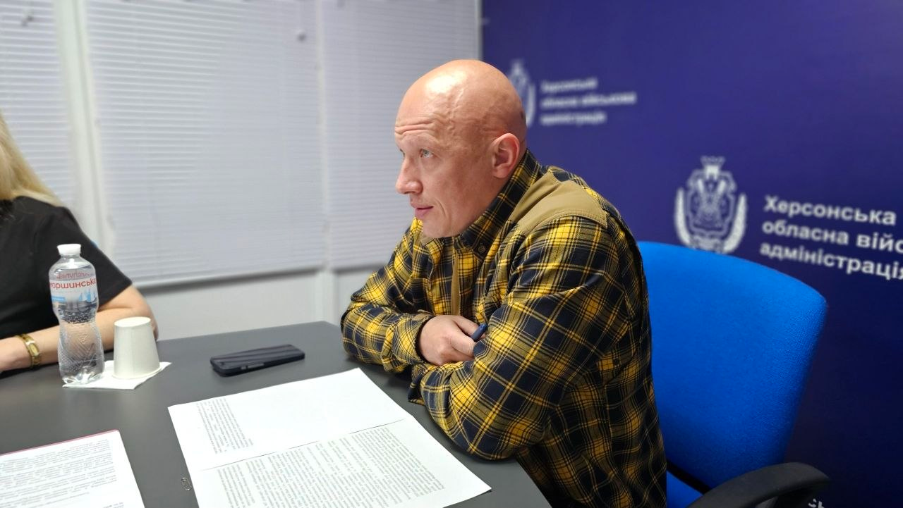 Чоловік у жовто-чорній карпованій сорочці сидить за столом під час наради чи офіційного виступу в кабінеті. Перед ним розкладені документи, а на синьому тлі позаду розміщено офіційні герби та написи державної установи.