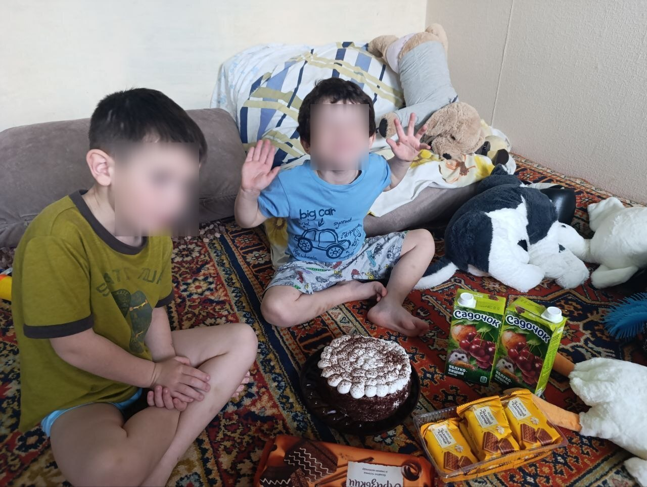 Двоє хлопчиків із розмитими обличчями сидять на візерунчастому килимі поруч із солодощами. Перед ними на підлозі стоїть шоколадний торт із білим кремом, два пакунки соку та печиво, а навколо розкладені м'які іграшки.
