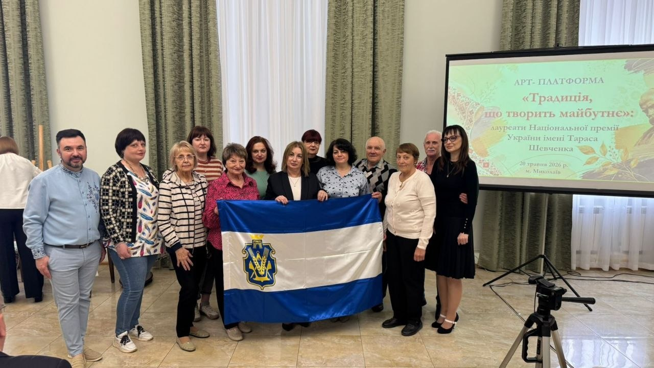 Групове фото учасників заходу в приміщенні біля великого проекційного екрана з написом «Арт-платформа «Традиція, що творить майбутнє»». Люди стоять разом та тримають перед собою офіційний синьо-білий прапор Херсонської області з гербом регіону у лівій частині.