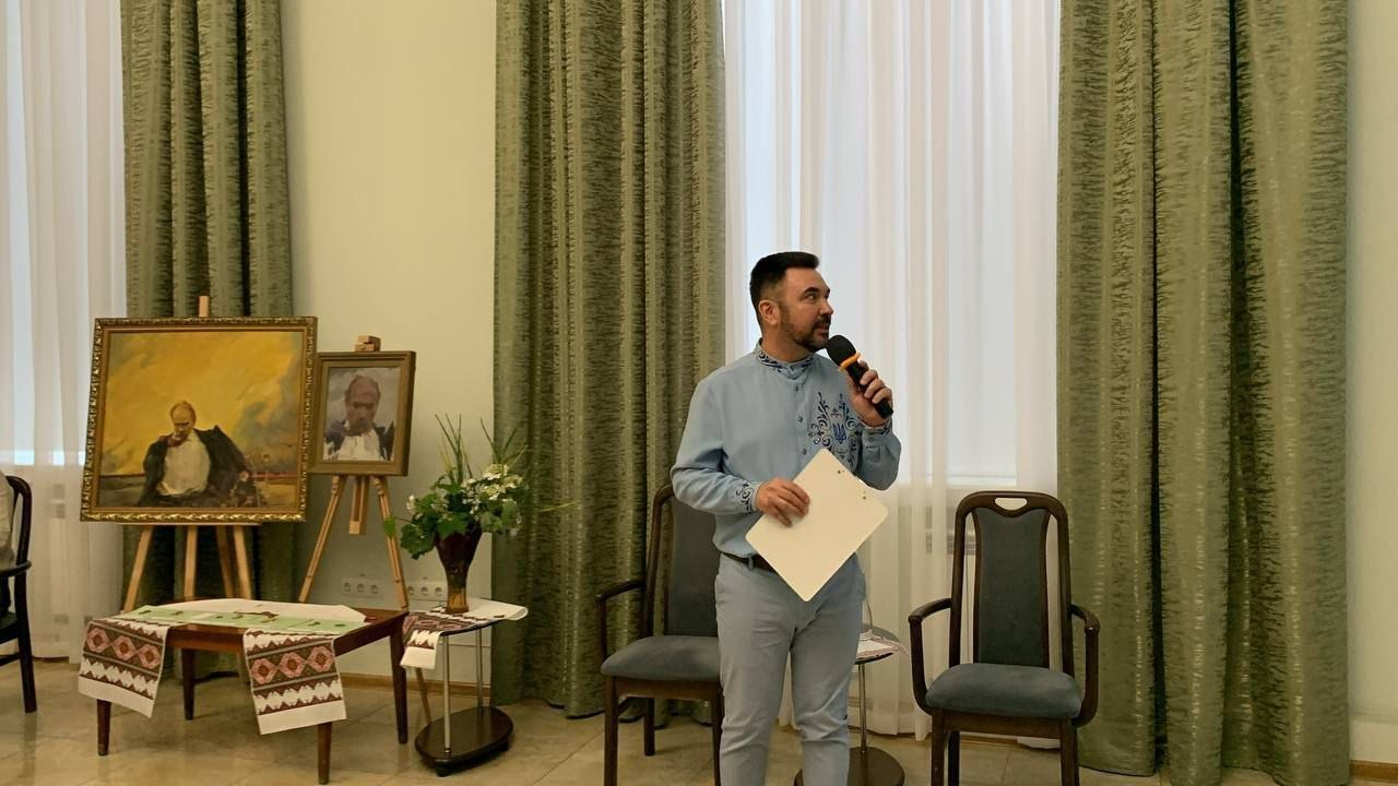 Чоловік у блакитній вишиванці виступає з мікрофоном та папкою в руках у залі з високими зеленими шторами. Ліворуч на мольбертах розміщено картини з портретами Тараса Шевченка, а поруч на столиках, застелених вишитими рушниками, стоїть ваза з квітами.