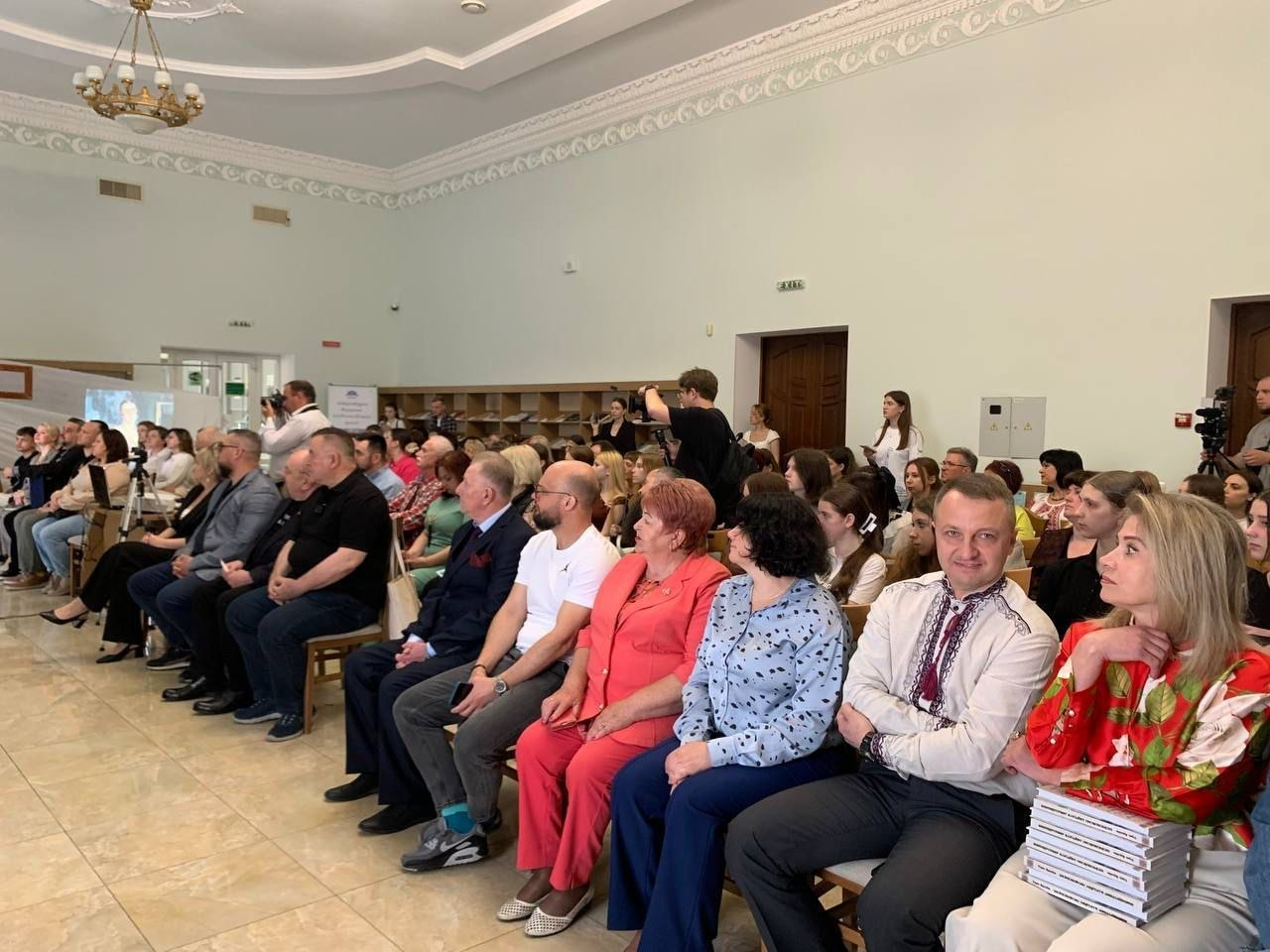 Фото заповненої глядацької зали під час офіційного заходу чи лекції. Люди різного віку сидять рядами на стільцях та уважно слухають виступ, дехто з присутніх одягнений у вишиванки. На задньому плані видно операторів із камерами, а біля дверей стоять інші слухачі.