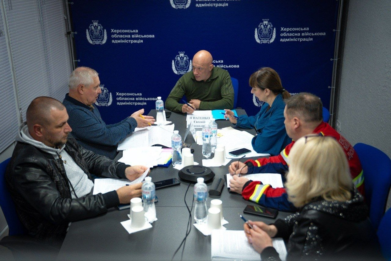 Група людей сидить за великим столом під час робочої наради чи засідання установи. Учасники обговорюють документи, перед ними розкладені папери та пляшки з водою, а на синьому тлі позаду багаторазово повторюються офіційні герби та написи установи.