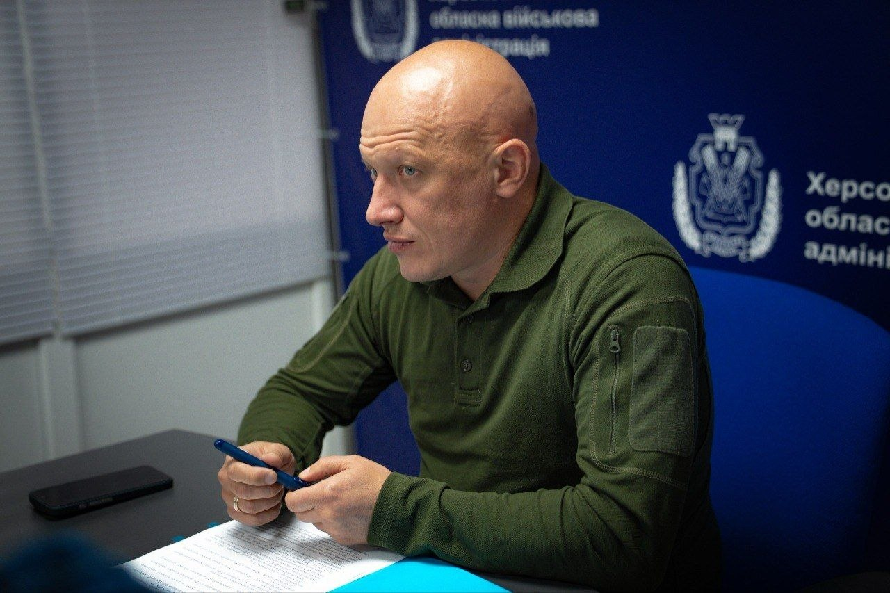 Чоловік у зеленій тактичній кофті сидить за столом під час наради чи офіційного виступу. Він тримає ручку в руках і дивиться вбік, перед ним лежать папери та смартфон, а на синьому тлі позаду видно герб та офіційні написи установи.