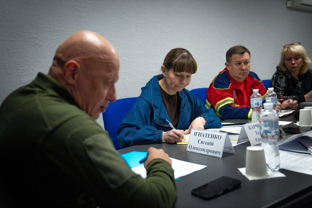 Чотири особи — двоє чоловіків та дві жінки — сидять за столом під час робочої наради й роблять записи в документах. Перед ними розкладені папери, канцелярія та пляшки з водою.