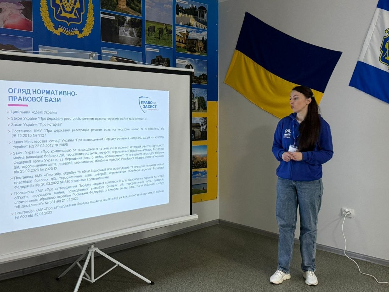 Жінка в синій кофті виступає біля проєкційного екрана зі слайдом правової презентації. Праворуч на стіні висить прапор України, а позаду розміщено інформаційний стенд.