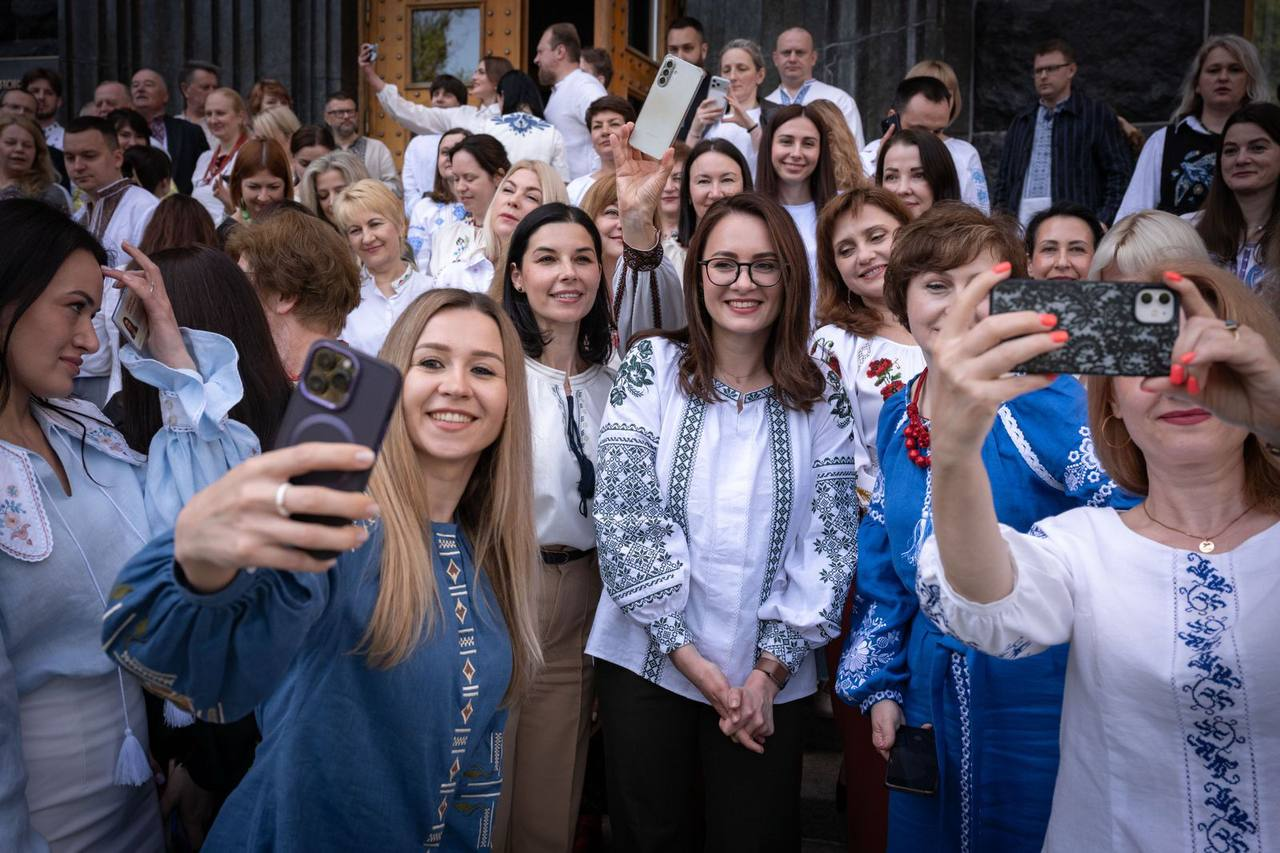 Групове фото великої кількості людей у традиційному українському одязі — вишиванках різних кольорів та орнаментів. На передньому плані жінки посміхаються та роблять селфі на мобільні телефони на тлі входу до будівлі.
