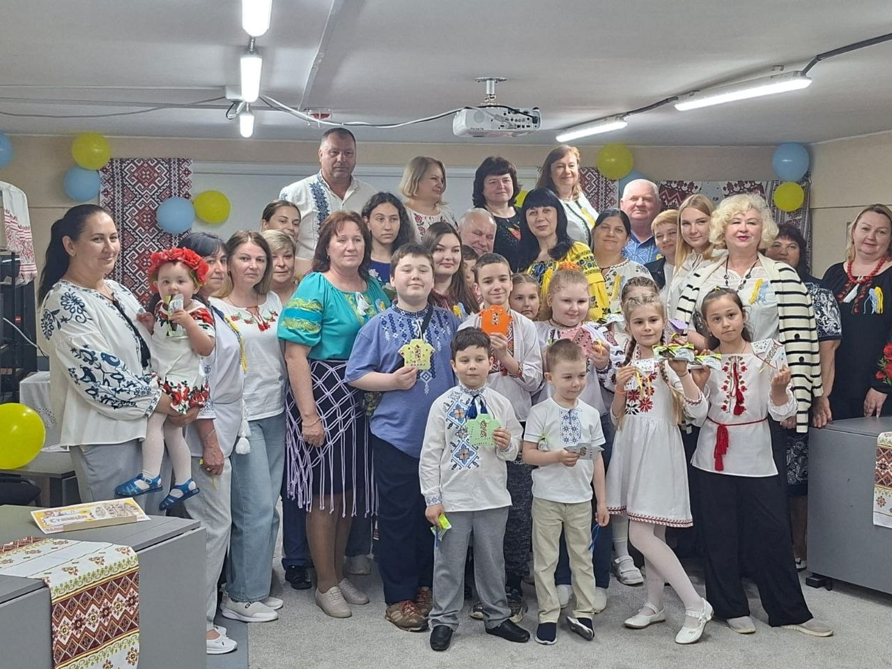 Групове фото дорослих та дітей у традиційному українському одязі та вишиванках у приміщенні. Вони стоять у кілька рядів, посміхаються та тримають у руках невеликі сувеніри й вироби. На задньому плані видно сині та жовті кульки, а також ткані рушники.