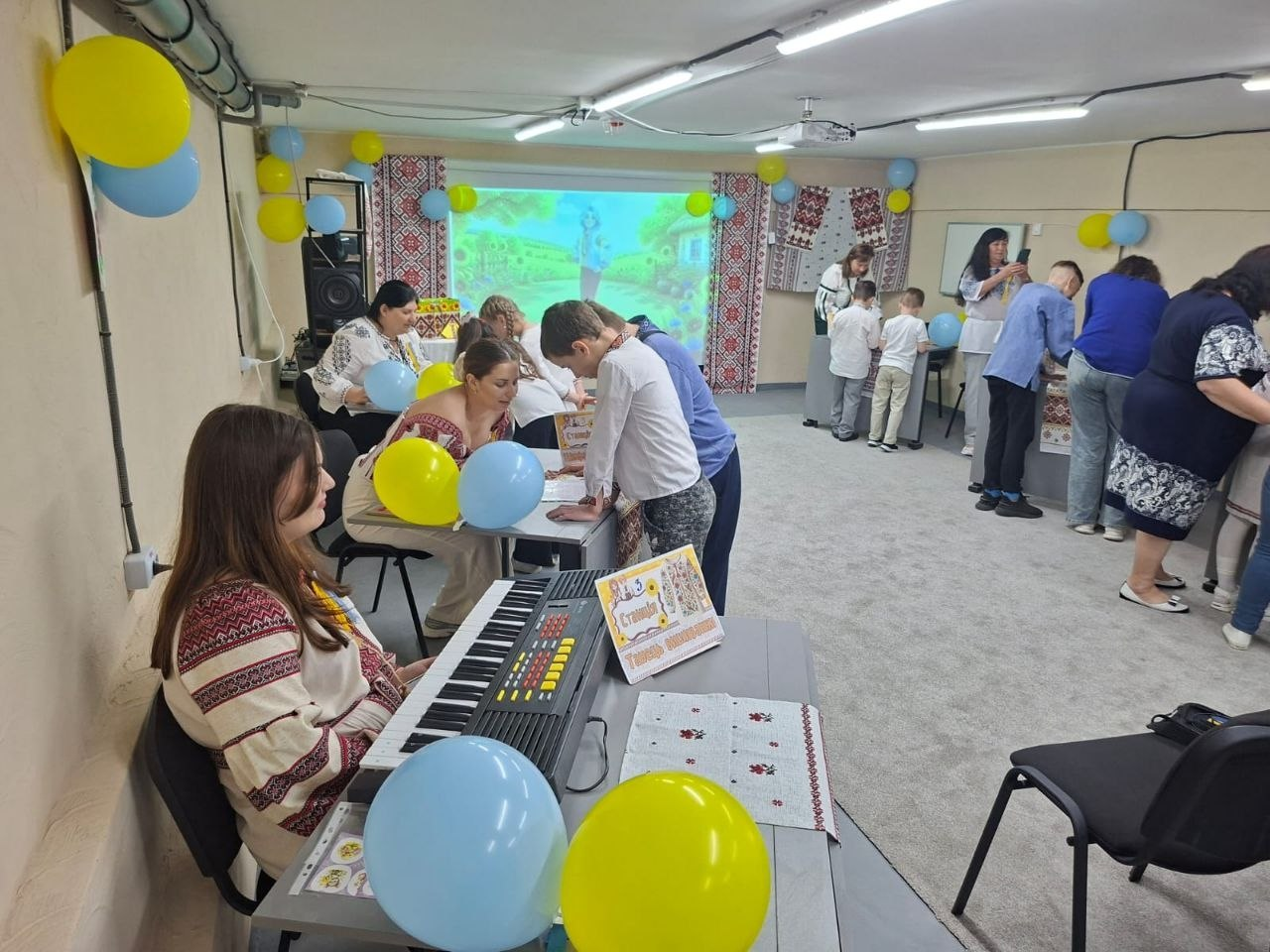 Спільний захід для дітей та дорослих у вишиванках, які виконують завдання за столами у світлому кабінеті. На передньому плані видно електронне піаніно, столи прикрашені синіми та жовтими повітряними кульками, а на задньому плані розміщено проєкторний екран та вишиті рушники.
