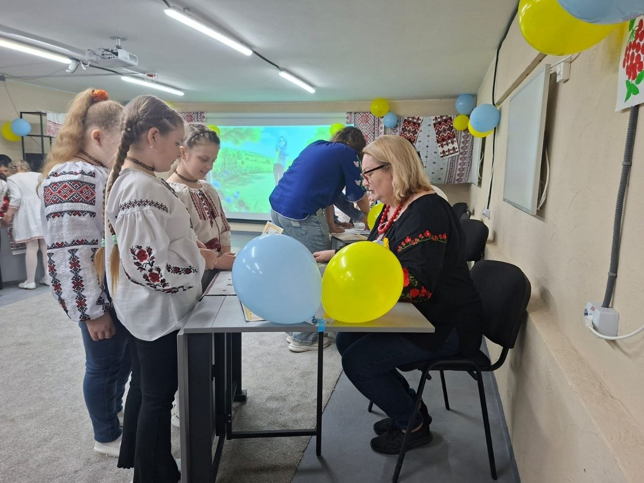 Три дівчинки у вишиванках стоять біля столу та спілкуються з дорослою жінкою в окулярах під час заходу в кабінеті. Стіл прикрашений синьою та жовтою кульками, а на задньому плані видно екран із зображенням, вишиті рушники та інших учасників.