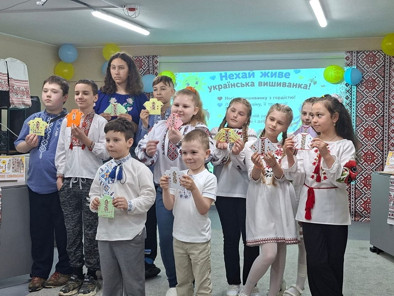 Група дітей у вишиванках стоїть у приміщенні та демонструє зроблені власноруч паперові сувеніри у формі сорочок. На задньому плані розміщено великий екран із написом «Нехай живе українська вишиванка!», повітряні кульки та вишиті рушники.