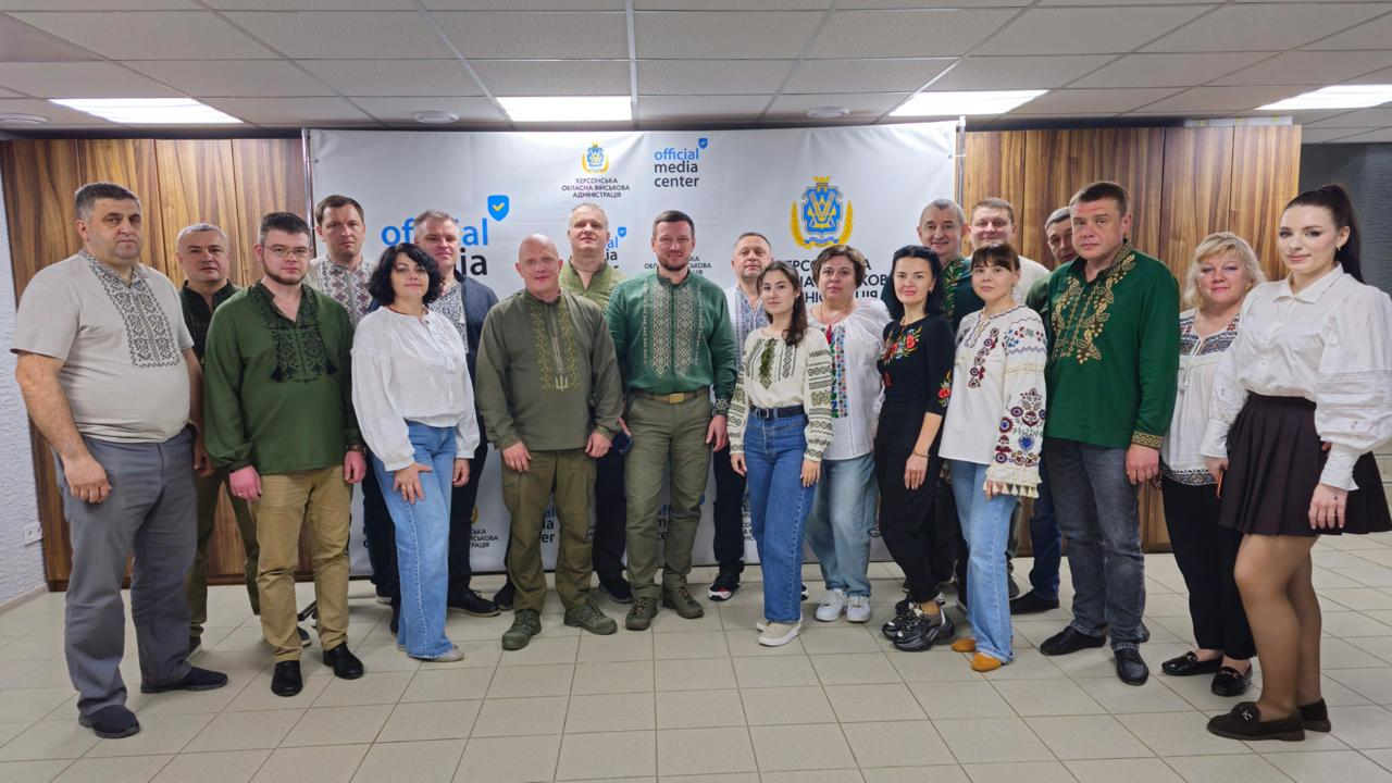 Групове фото чоловіків та жінок у різноманітних вишиванках, які стоять у два ряди у приміщенні. Вони позують на тлі білого банера з написами «official media center» та гербом області, а також дерев'яної стіни.