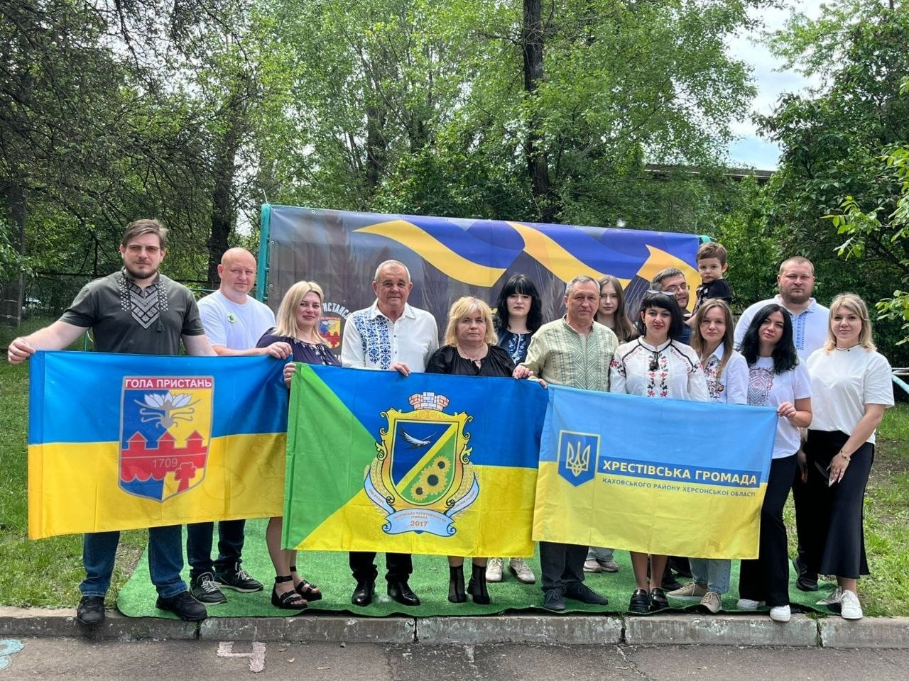 Групове фото людей на тлі дерев, які тримають у руках прапори Голої Пристані та Хрестівської громади Каховського району Херсонської області, а також ще один синьо-жовто-зелений стяг із символікою. Деякі учасники вбрані у традиційні українські вишиванки.