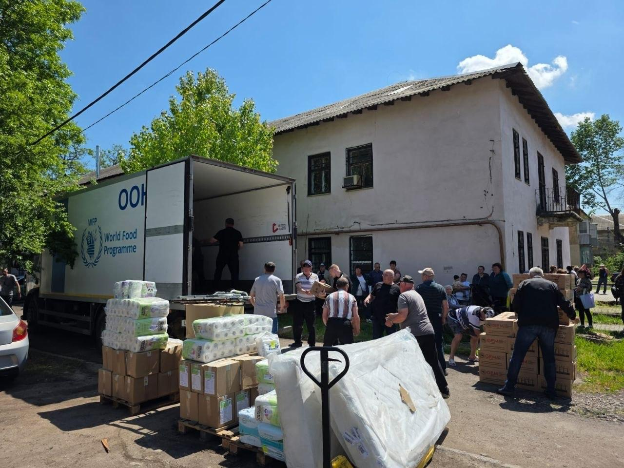 Процес розвантаження гуманітарної допомоги з вантажівки Всесвітньої продовольчої програми (WFP) біля двоповерхової будівлі. Група людей переносить коробки та упаковки з гігієнічними засобами, які складені на дерев'яних піддонах просто неба.