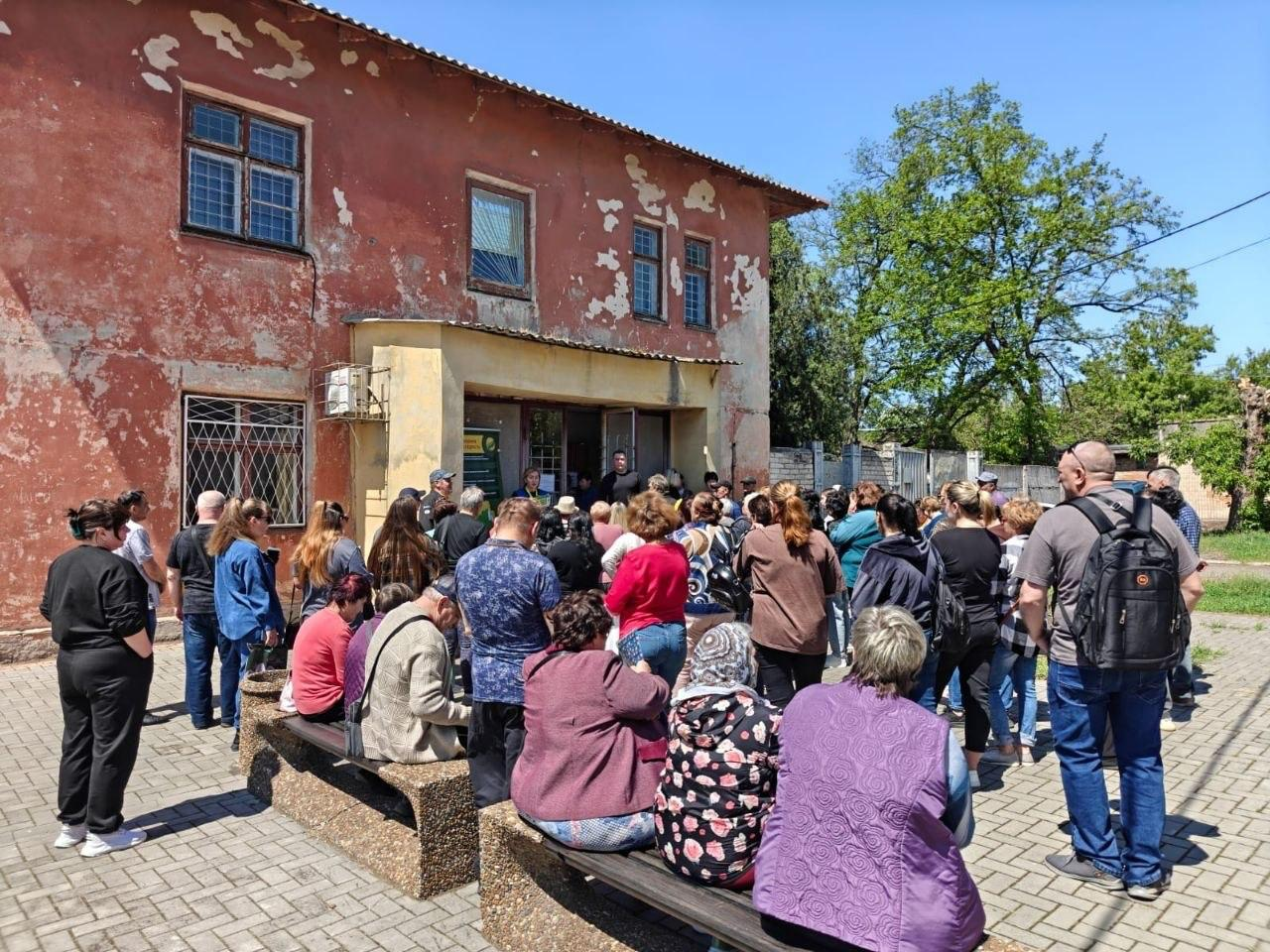 Велике скупчення людей біля входу до двоповерхової адміністративної будівлі з полущеною рожевою фарбою на стінах. Мешканці стоять у черзі та на лавках просто неба, очікуючи на видачу допомоги в сонячний день.