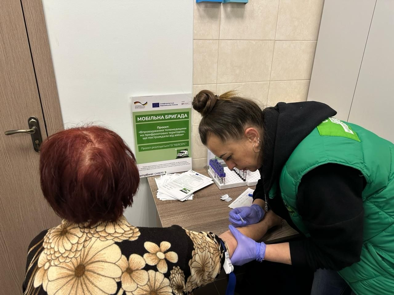 Медична працівниця у зеленому жилеті та гумових рукавичках бере кров із вени у жінки похилого віку в кабінеті. На столі поруч розміщено пробірки, бланки та інформаційний плакат мобільної бригади про проєкт із телемедицини.
